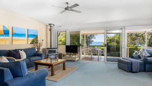A spacious living room with a large sofa, a coffee table, and a TV. There is a fireplace and a balcony with a table and chairs.