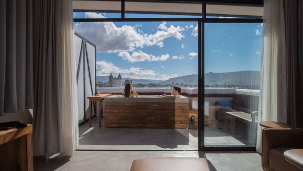 A modern room with a large glass window, a wooden bench, and a view of a cityscape. - SUITE DAWN (Oaxaca de Juárez)