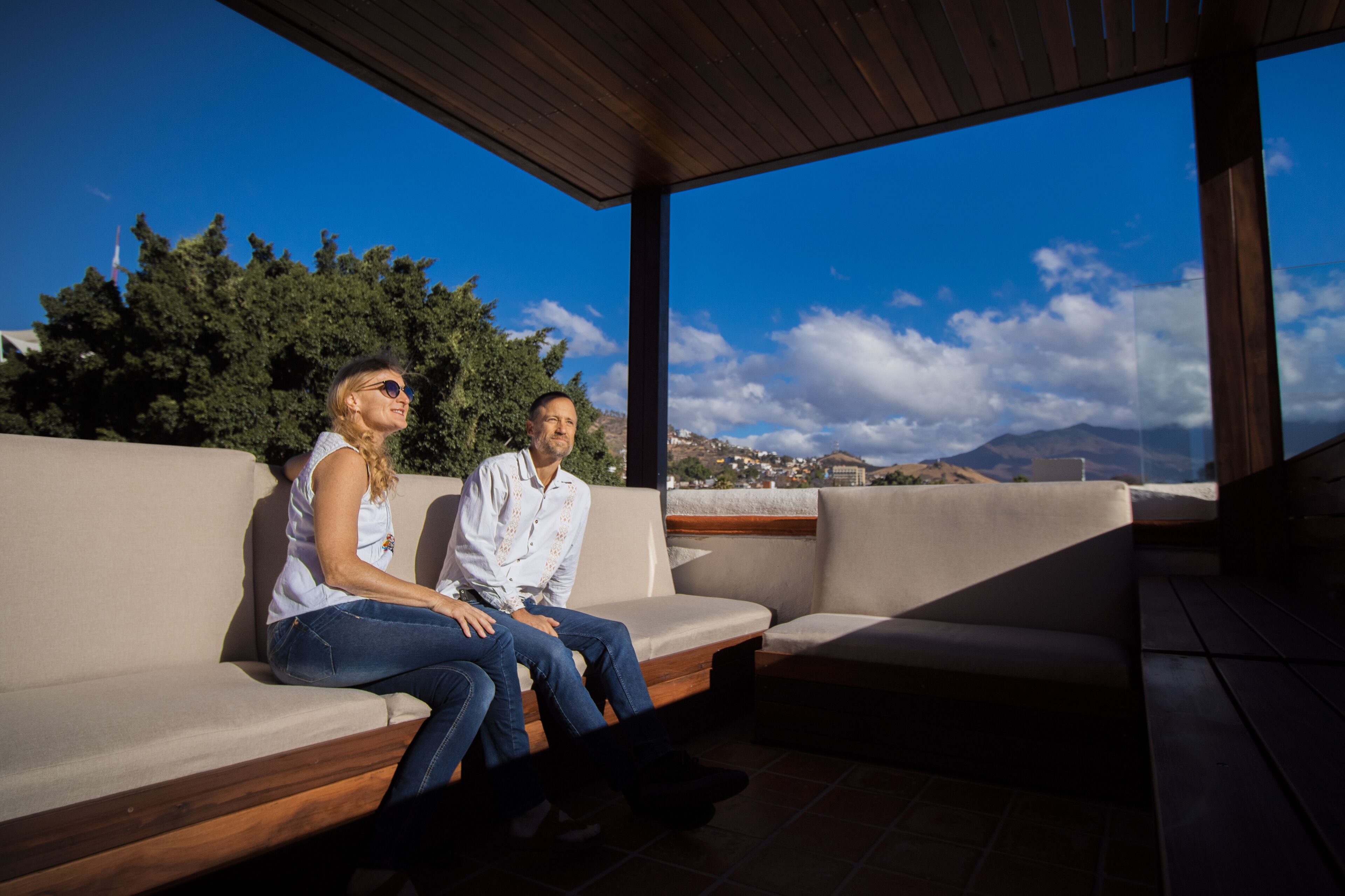 A couple sitting on a beige sofa with a wooden ceiling and large windows overlooking a mountainous landscape.