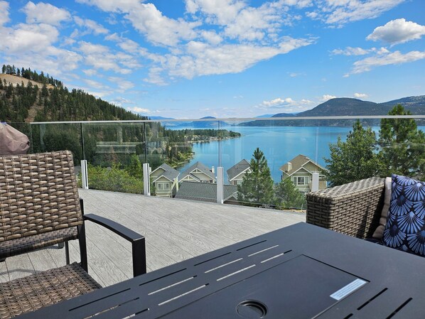 A terrace with wicker furniture, a glass balustrade, and a view of a lake and mountains.