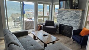 A modern living room with a stone fireplace, grey sofas, and a large window overlooking a scenic view. - FULLY RENOVATED LAKEVIEW COTTAGE / PET FRIENDLY - LA CASA (Kelowna)