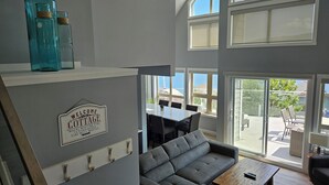 A modern living room with a grey sectional sofa, a wooden coffee table, and a dining area with a table and chairs. There are large windows providing natural light and a view of the outside. - FULLY RENOVATED LAKEVIEW COTTAGE / PET FRIENDLY - LA CASA (Kelowna)