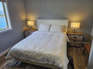 A neatly made bed with white linens, two bedside tables with lamps, and a view of the outdoors through a window.