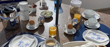 A breakfast table set with plates, cups, and a basket of bread, overlooking a coastal view.
