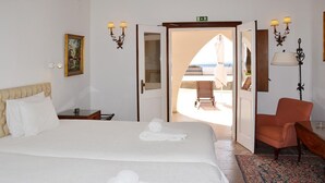 Ett hotellrum med två sängar, en sittgrupp med en stol och ett litet bord, och utsikt över stranden genom en öppen dörr.