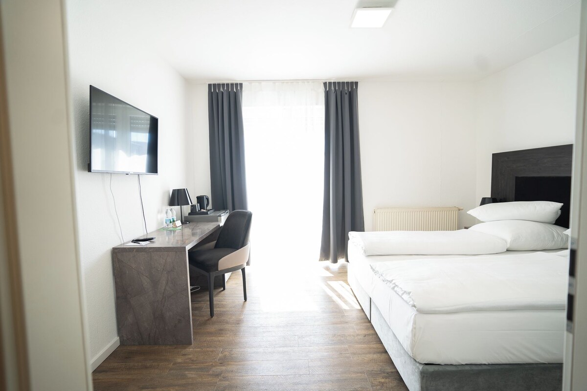A hotel room with a bed, a desk with a chair, a TV mounted on the wall, and a window with curtains.