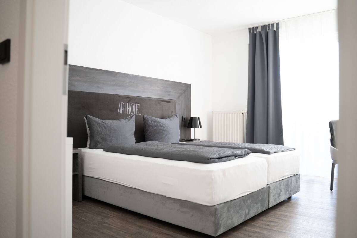 Una habitación de hotel moderna con una cama grande, ropa de cama gris, un cabecero de madera, una mesita de noche con lámpara y vistas al exterior.