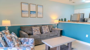 Ein Wohnzimmer mit einer Couch, zwei gemusterten Sesseln, einem Couchtisch und einer Küchenzeile mit Kühlschrank und Schränken.