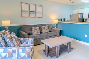 A living room with a sofa, two patterned armchairs, a coffee table, and a kitchen area with a refrigerator and cabinets.
