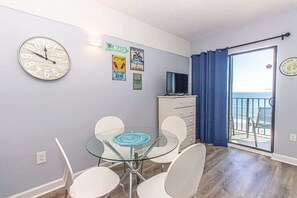Una habitación con una mesa de vidrio, sillas blancas, un reloj, un televisor y vista al océano desde un balcón.