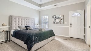 Quarto com cama de cabeceira estofada, ventilador de teto, janela com persianas, porta e quadro pendurado na parede.