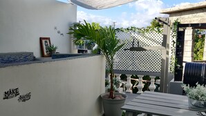 En terrasse med et bord og stole, en potteplante samt en væg med et indrammet billede og et motiverende citat.