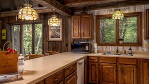 Une cuisine rustique avec des armoires en bois, un four à micro-ondes et un évier. Deux luminaires sont suspendus au plafond, et une fenêtre offre une vue sur les arbres. Un sac de bienvenue est posé sur le comptoir.