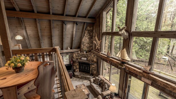 L’intérieur d’un chalet rustique, avec une cheminée en pierre, des meubles en bois et de grandes fenêtres donnant sur la forêt.