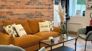 Un salon avec un mur en briques, un canapé orné de coussins sur lesquels est écrit le mot « MAISON », une table basse et un fauteuil.