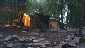 A rustic cabin surrounded by a stone wall and trees, with a small porch illuminated by warm light.