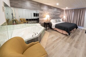 A modern hotel room with a large bathtub, a bed with black bedding, a bedside table with a lamp, and a large mirror.