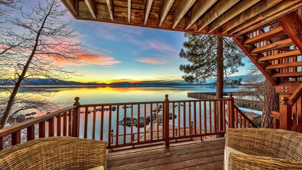 A wooden deck with wicker chairs overlooking a calm lake at sunset.