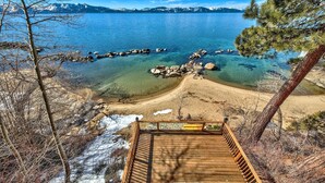 A wooden deck with a bench and a view of a lake with clear water, rocks, and mountains in the distance.