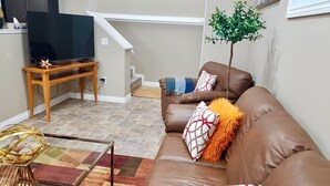 A living room with a brown leather sofa, a glass coffee table, a television on a wooden stand, and a staircase leading to an upper floor. - ❤️Executive Suite❤️By 2 Shopping Centres, Costco ... (Grande Prairie)