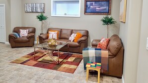 A living room with a brown leather sofa set, a glass coffee table, and a colorful rug. There are two potted plants, a framed picture on the wall, and a window with blinds. - ❤️Executive Suite❤️By 2 Shopping Centres, Costco ... (Grande Prairie)