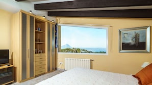 Une chambre avec un grand lit, une armoire, un téléviseur, un radiateur et une fenêtre donnant sur une vue imprenable sur la montagne et la mer.