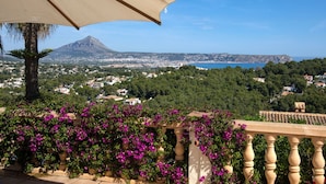 Un balcon donnant sur une ville côtière, avec une montagne en arrière-plan et des fleurs violettes au premier plan.