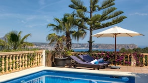 Un espace piscine équipé de chaises longues, d’un parasol et offrant une vue sur la mer et les collines.