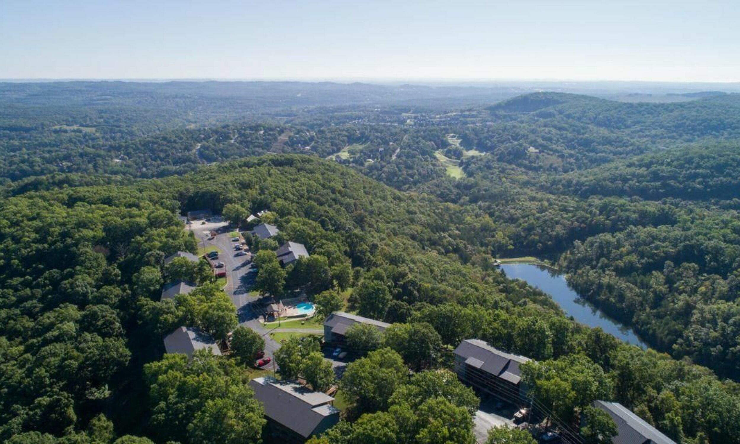 An aerial view of a residential area with houses, a swimming pool, and a lake surrounded by dense forest.