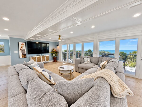 A spacious living room with a large sectional sofa, a flat-screen TV, and a balcony with a view of the ocean.