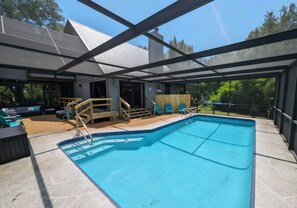 A covered outdoor swimming pool with a wooden deck, seating area, and a view of the surrounding landscape.