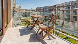 Ein Balkon mit Holztisch und -stühlen, der Blick auf moderne Gebäude und einen darunterliegenden Garten gewährt.