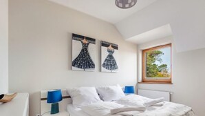 Un dormitorio con cama, dos lámparas fijadas a la pared, una ventana con vistas a árboles y dos cuadros abstractos.