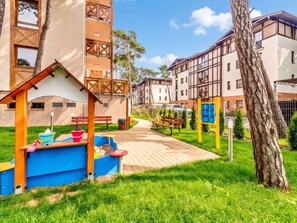 A playground with a slide and sandpit, surrounded by a grassy area and a pathway leading to a building with multiple windows.