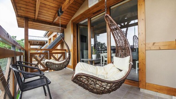 Un balcón con dos sillas de mimbre y una hamaca colgante, vigas de madera y un ventanal de vidrio que da a un espacio al aire libre.