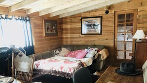 A rustic cabin interior with a wooden ceiling, a sofa bed, a dining table, a chair, and a cowhide rug. - Vacation with the Horses in our Ranch House! Right off Hwy 395 (Inyokern)