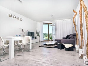 Een moderne woonkamer met een witte eethoek, een grijze bank en een groot raam met gordijnen.