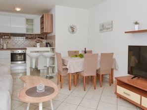 Una cocina moderna con armarios blancos y un horno empotrado. Hay un comedor con una mesa y sillas, y un televisor sobre un mueble de madera.