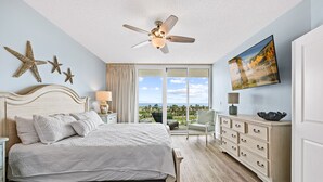 A bedroom with a bed, a ceiling fan, a nightstand, a dresser, a chair, and a large window with a view.