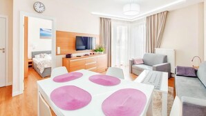 A modern hotel room with a dining area, a living area with a sofa and armchair, a TV, and a clock on the wall.