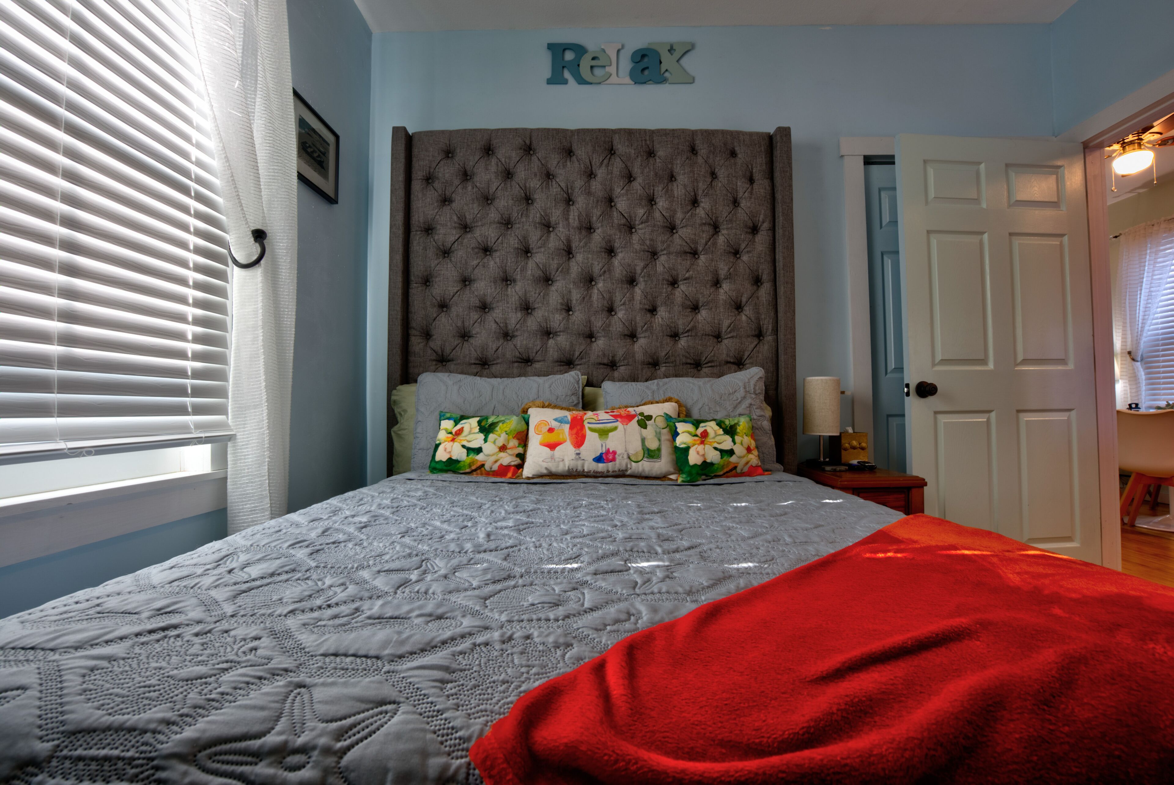 A bedroom with a tufted headboard, a bed with patterned bedding, a red throw blanket, and a 'Relax' sign on the wall.