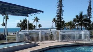 A clear blue pool with two people swimming, surrounded by palm trees and a covered area.