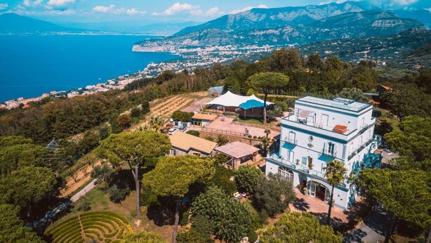 Un edificio grande de varios pisos con una fachada blanca destacada y detalles azules, rodeado de exuberante follaje y un paisaje costero.