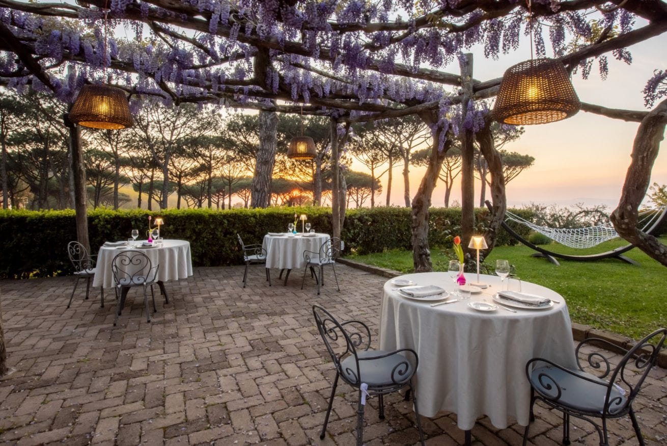An outdoor dining area with round tables and chairs, covered by a pergola with purple wisteria, and a hammock in the background.