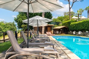 A poolside area with lounge chairs and umbrellas, a stone pathway, and a small building in the background.