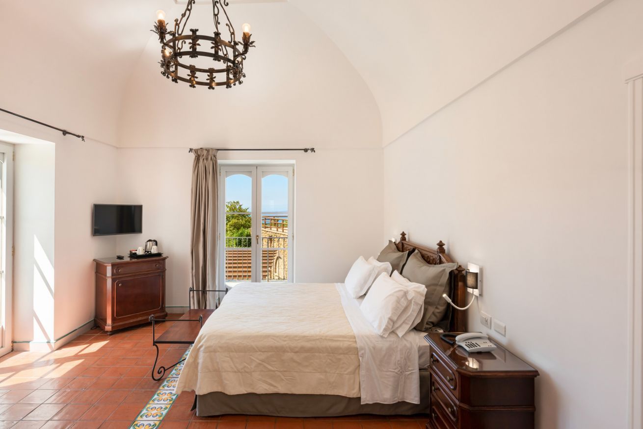 A bedroom with a bed, a desk with a telephone, a television on a cabinet, and a chandelier.