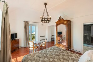 Ein Hotelzimmer mit Bett, Holzkommode, Fernseher, Esstisch mit Stühlen, einer Kristallleuchtre, einem großen Spiegel und einer Glastür zu einem Balkon.