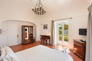 A bedroom with a large bed, a desk, a chair, a wardrobe, a television, and a sliding glass door leading to a balcony.
