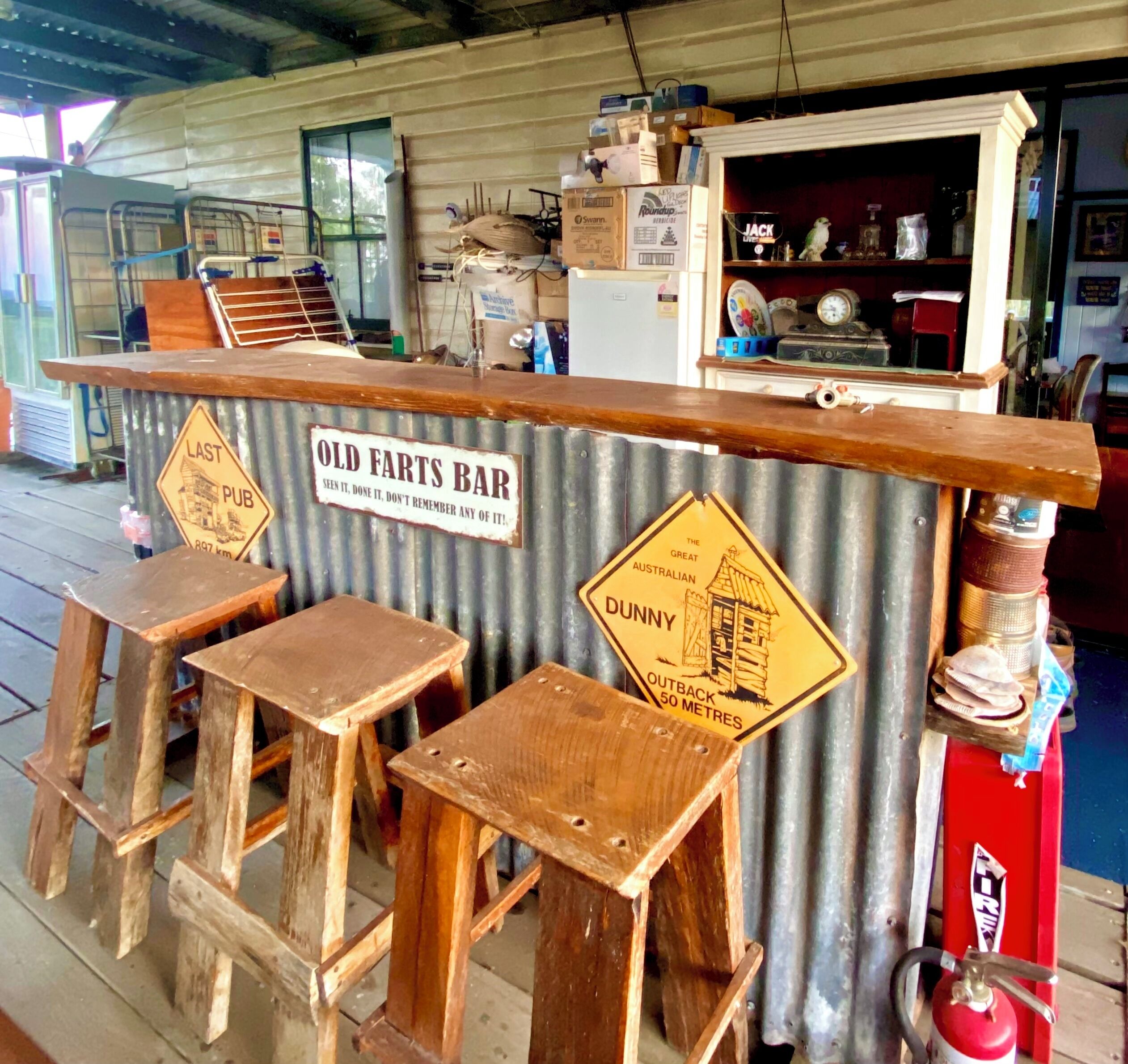 Een rustieke bar met houten krukken en een golfplaten achtergrond, met borden zoals 'Old Farts Bar' en 'Dunny Outback 50 Metres'.