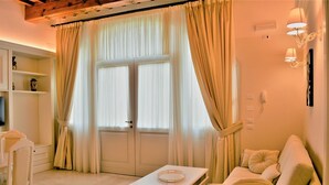 Ein Zimmer mit einem weißen Sofa, einem kleinen Tisch und einem Fenster mit beige farbenen Vorhängen. Es gibt ein weißes Regal mit Dekorationen sowie einen weißen Sessel.